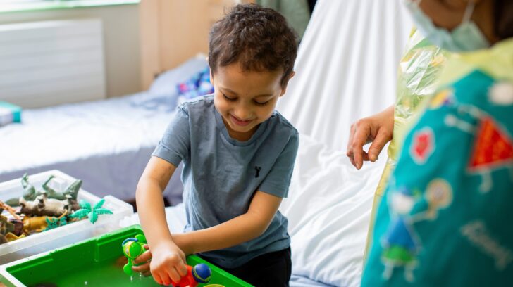 A young boy plays with toys at a bedside tray while a staff member stands nearby, supporting him.